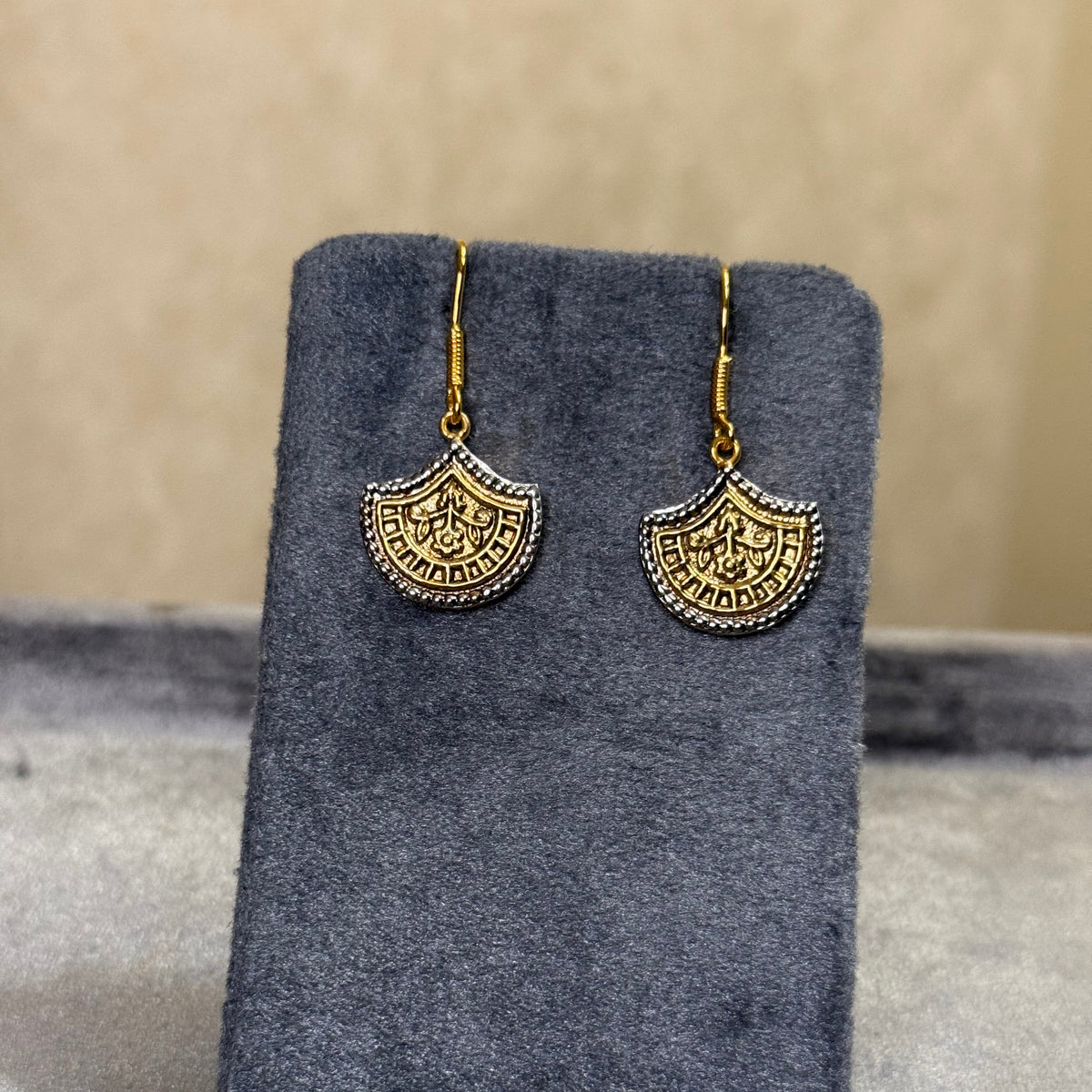 A pair of gold-toned dangle earrings with ornate circular cutout details, displayed on a dark gray velvet jewelry card against a neutral gray background.
