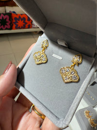 Gold-toned heart-shaped dangle earrings with clear rhinestone embellishments, displayed inside a gray velvet jewelry box held by a hand.
